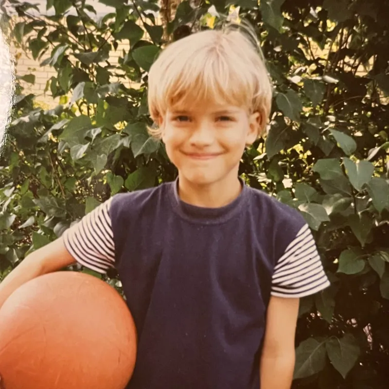 Young Anna Ring as a child in Jutland, standing in a garden with dappled sunlight, holding an orange ball, wearing a navy t-shirt with striped sleeves.