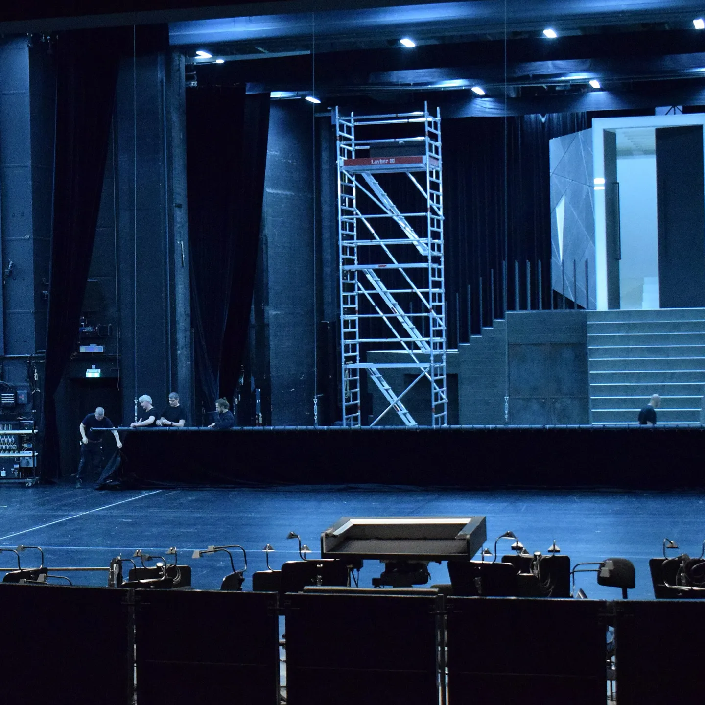 Opera stage in preparation, showing the conductor’s podium in the foreground and a set design with scaffolding under stage lights.